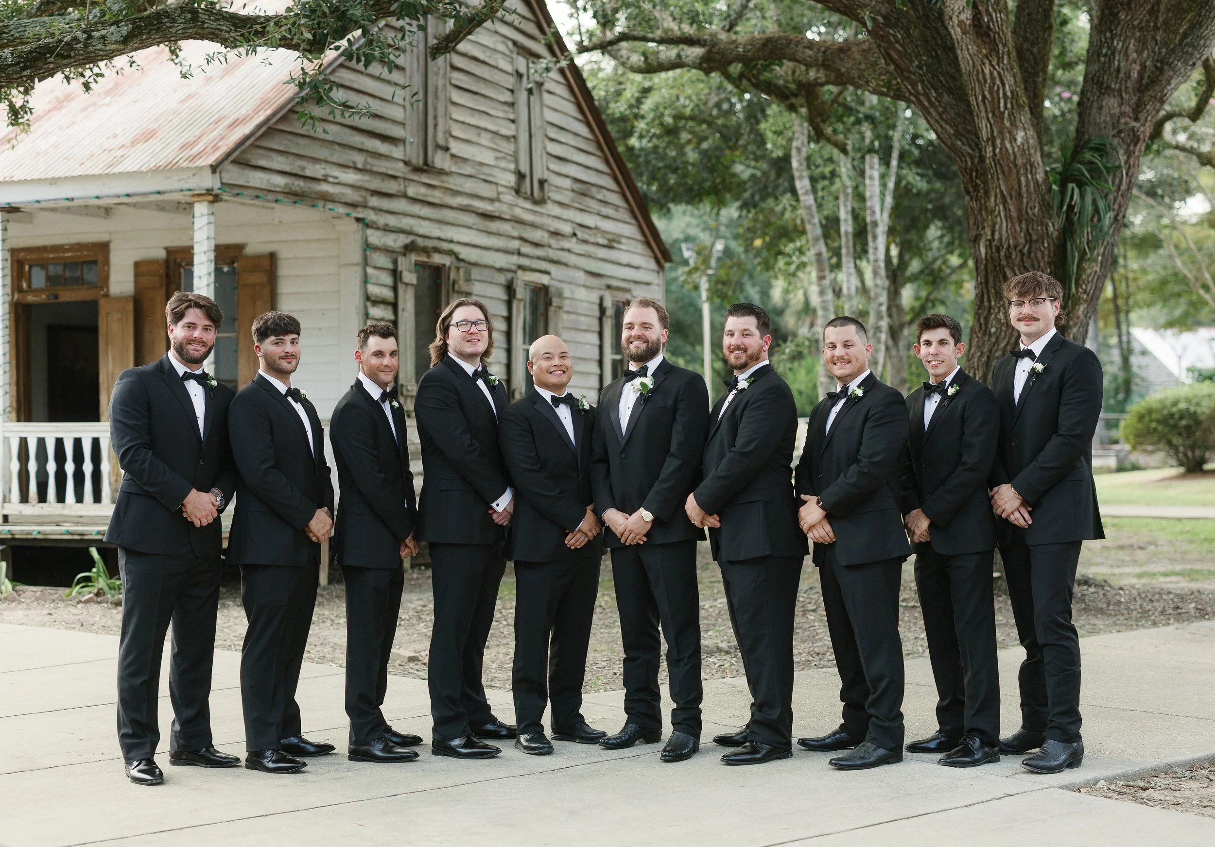 A group of nine men in tuxedos standing outdoors in front of a rustic wooden house and large trees, smiling for a photo.