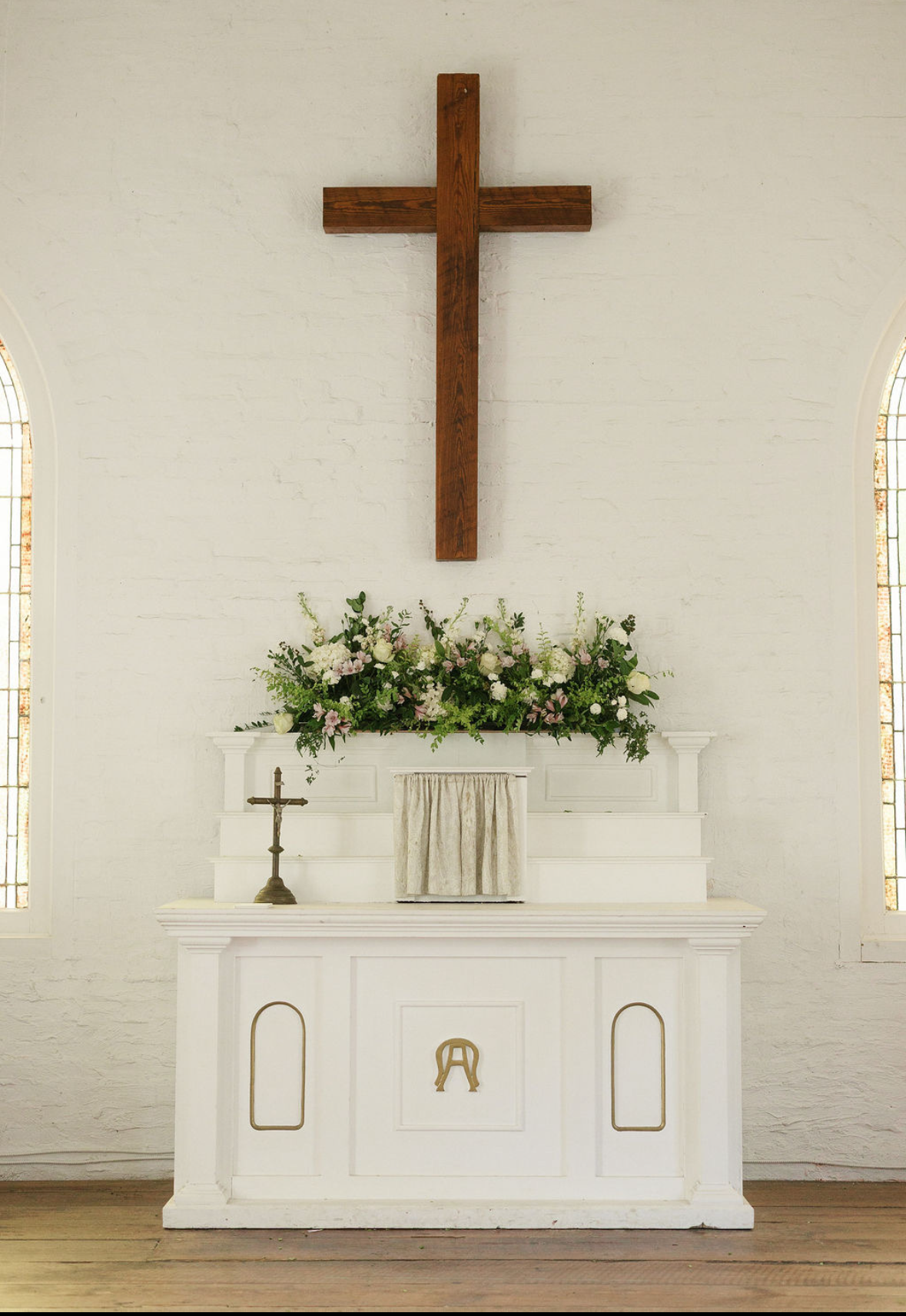 An altar inside a church with a large wooden cross, floral arrangement, small cross, and white cloth on the white altar table, flanked by tall narrow stained glass windows.
