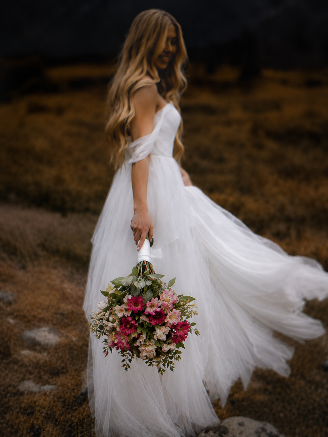 A woman in a white wedding dress holding a bouquet of pink and white flowers outdoors on a rocky terrain, with her long wavy hair flowing.