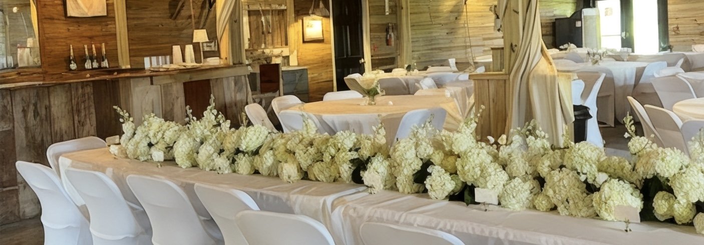 A wedding reception setup in a rustic wooden venue with long tables decorated with white tablecloths and a large floral centerpiece made of white hydrangeas, with chairs draped in white covers.