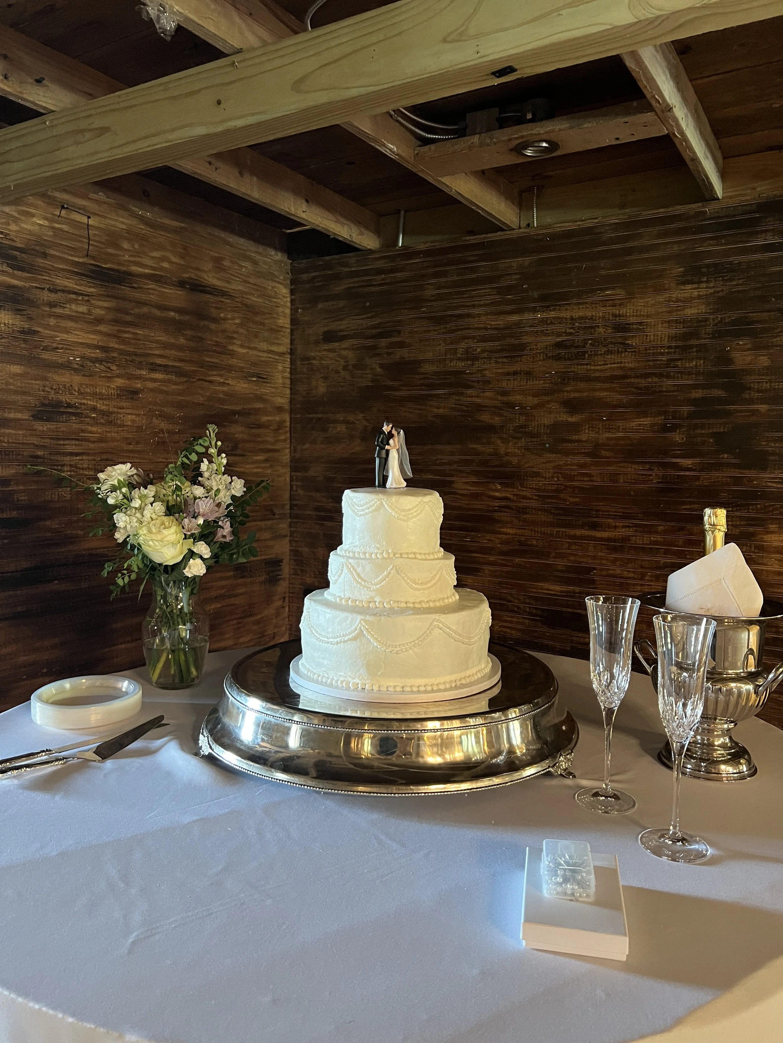 Wedding cake with a bride and groom topper, placed on a table with floral arrangements, glassware, and utensils, against a wooden wall background.