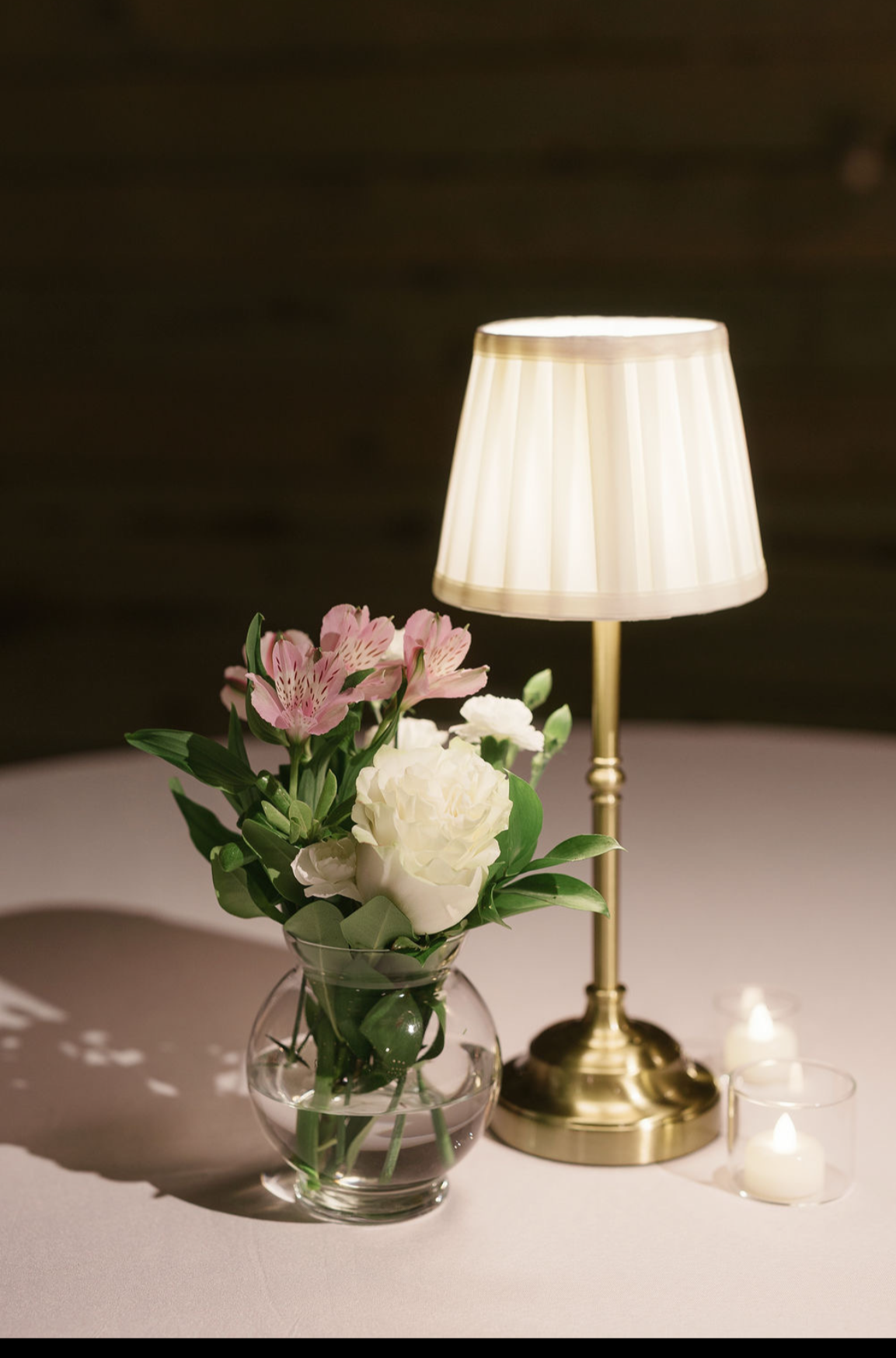 A table with a glass vase of pink and white flowers, a brass lamp with a white lampshade, and small lit candles, on a white tablecloth.