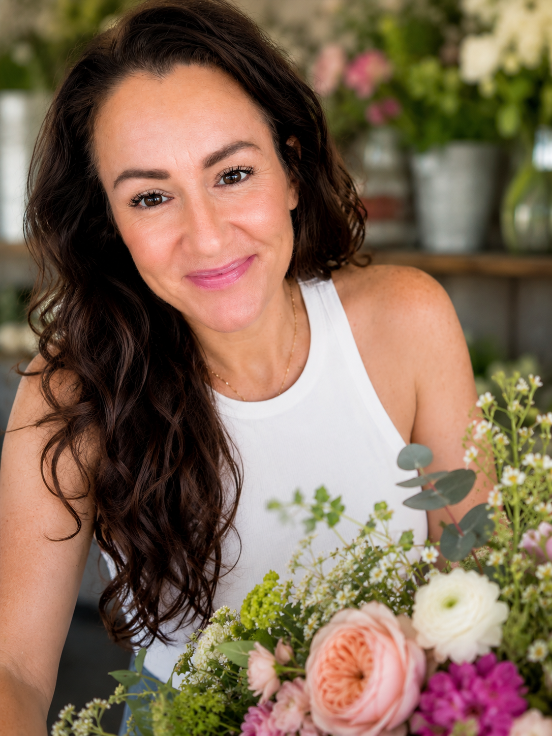 A woman with long, curly brown hair, smiling, sitting among a colorful flower arrangement with pink, white, and green flowers.