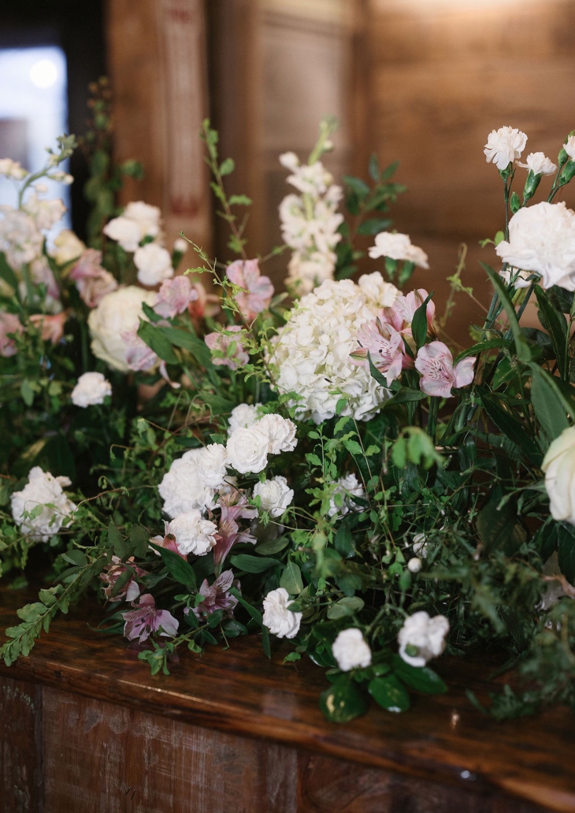 A floral arrangement of white and light pink flowers with green foliage on a wooden surface inside a room with wooden walls.