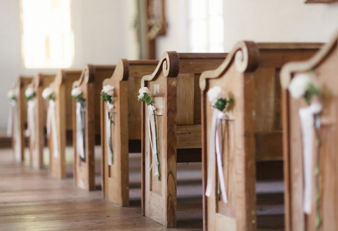 Wooden church pews decorated with white flowers and ribbons for a wedding ceremony.