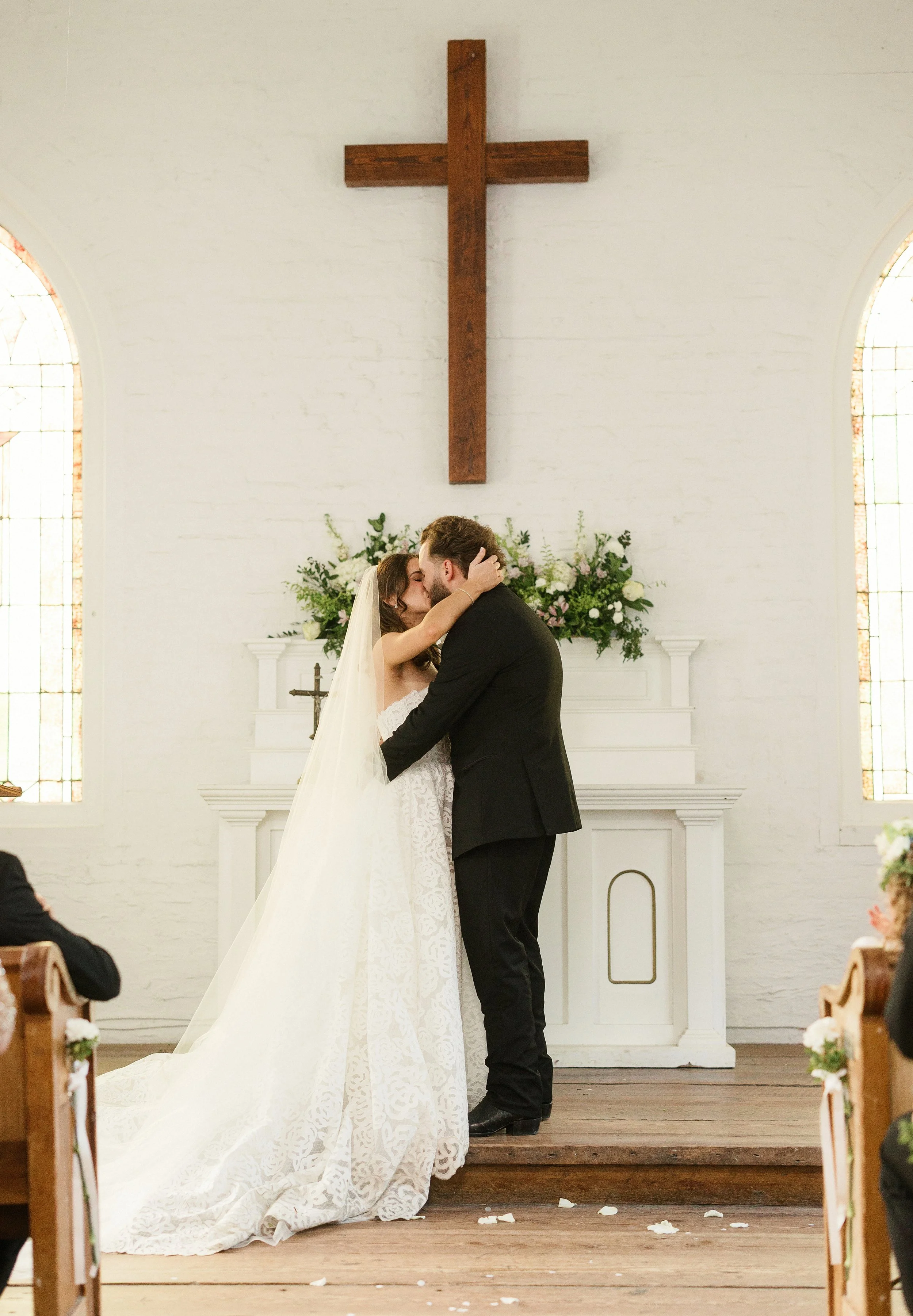 A bride and groom kiss during their wedding ceremony inside a church with a white brick wall, stained glass windows, a wooden cross, and floral arrangements.