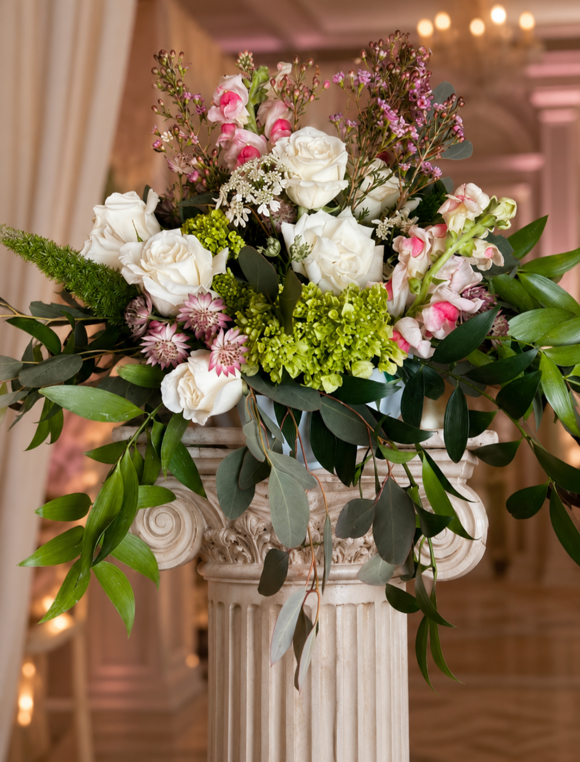 Elegant floral arrangement of white roses, pink snapdragons, and various greenery in a classical white urn on a pedestal.