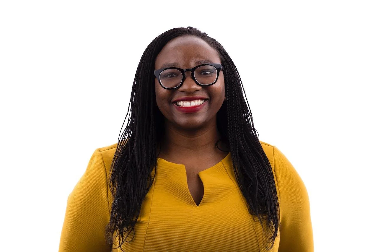 Dr. Dendere posing for a headshot, wearing a yellow dress, and glasses with black frames