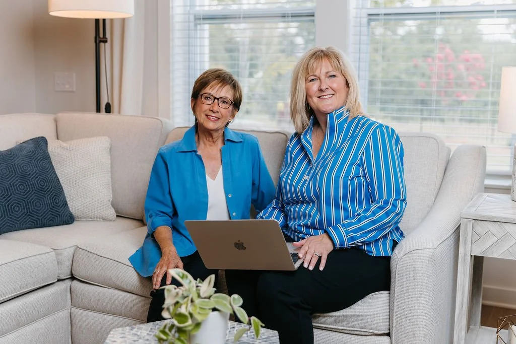 Two women, one elderly and one middle-aged, sitting on a beige couch with a laptop between them in a well-lit living room with window blinds, patterned pillows, and houseplants.