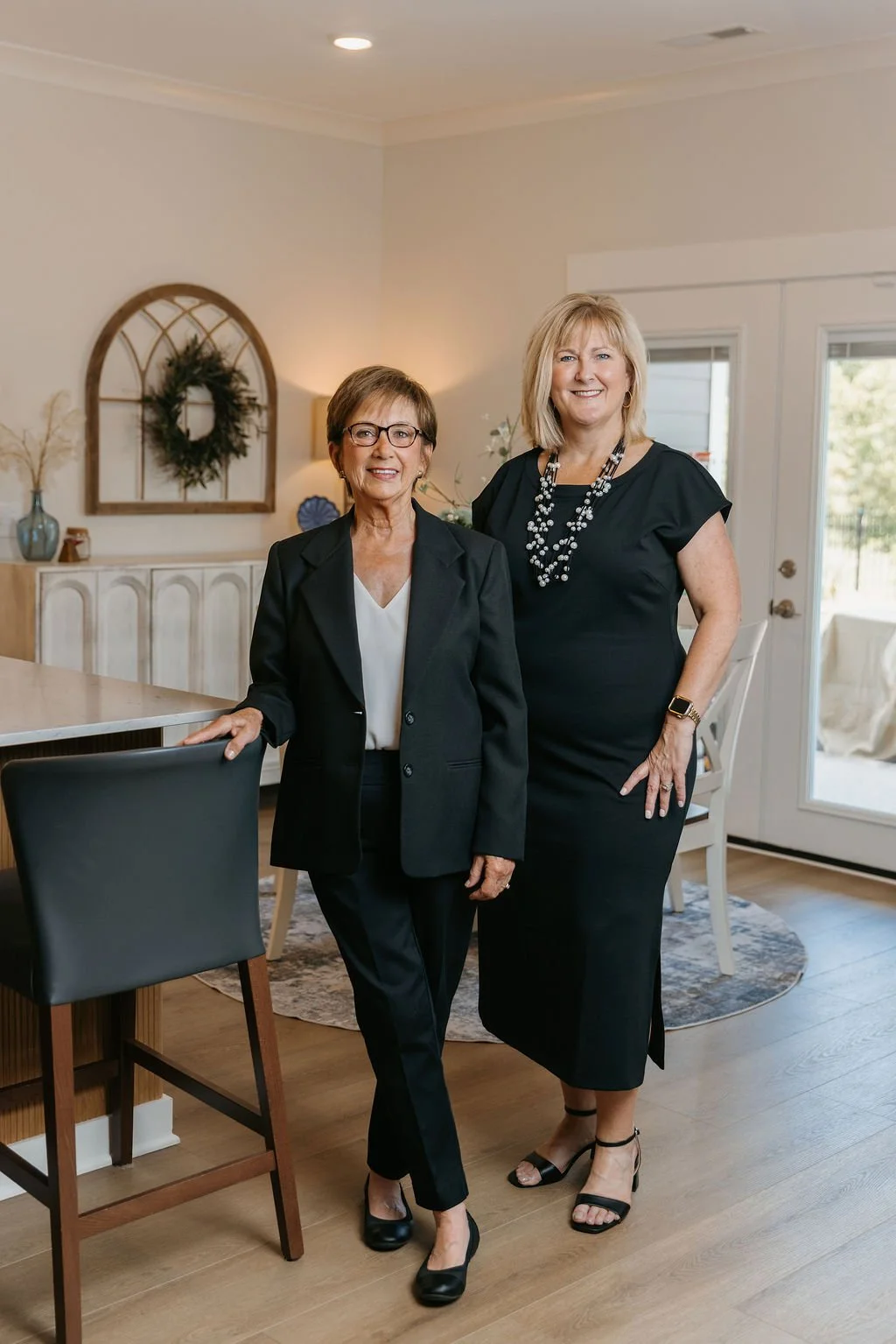 Two women standing in a living room, dressed in black professional attire, smiling at the camera.