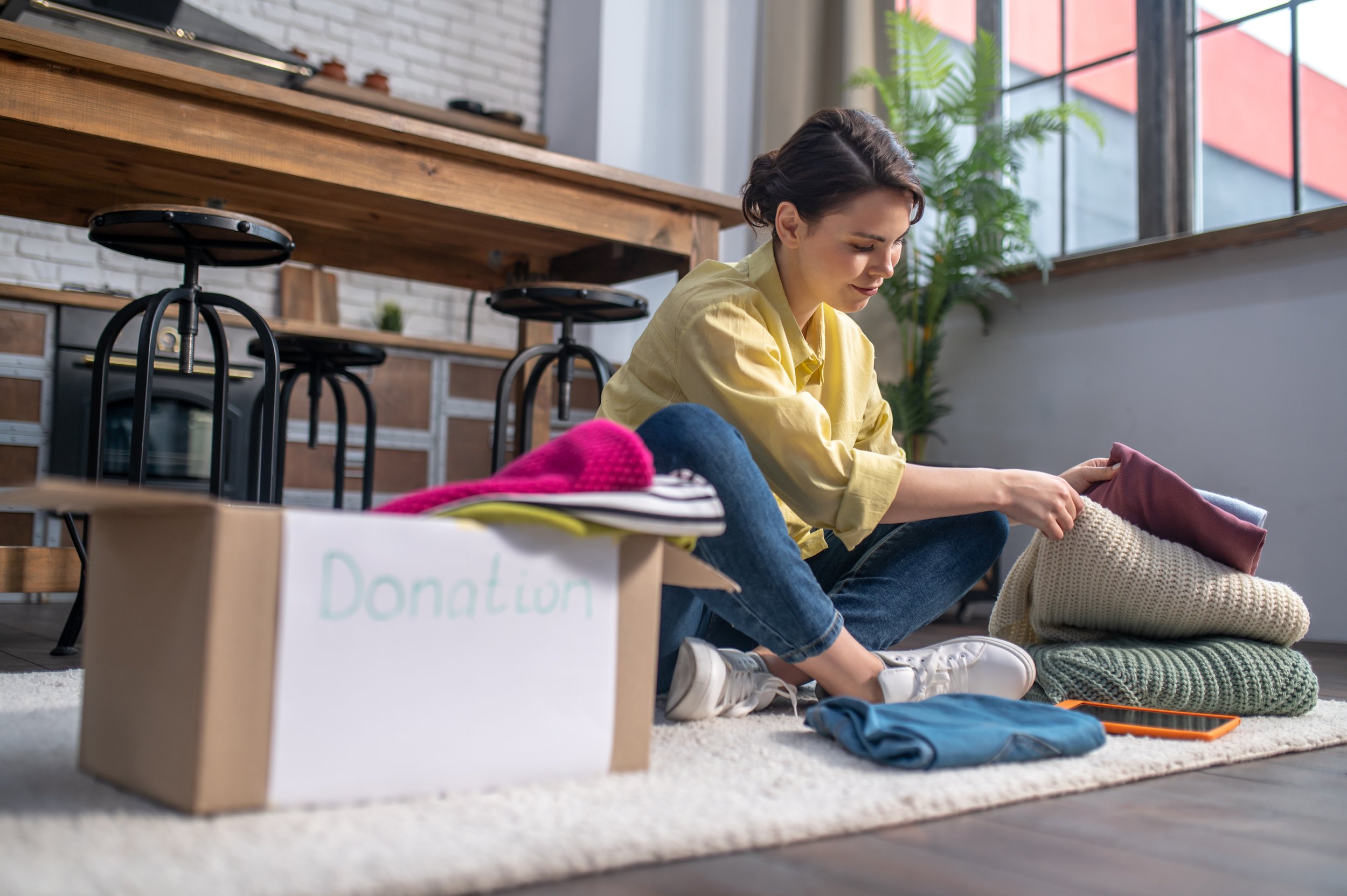 Woman organizing clothing and blanket donations in a loft-style room with large windows and potted plants.