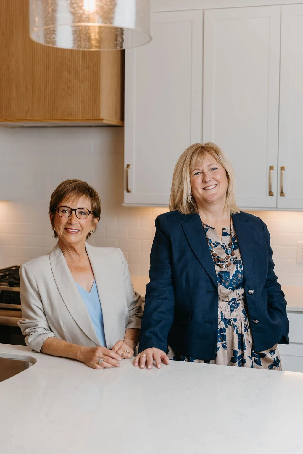 Two women smiling and standing in a modern kitchen, one in a white blazer and glasses, the other in a navy blazer and patterned dress.