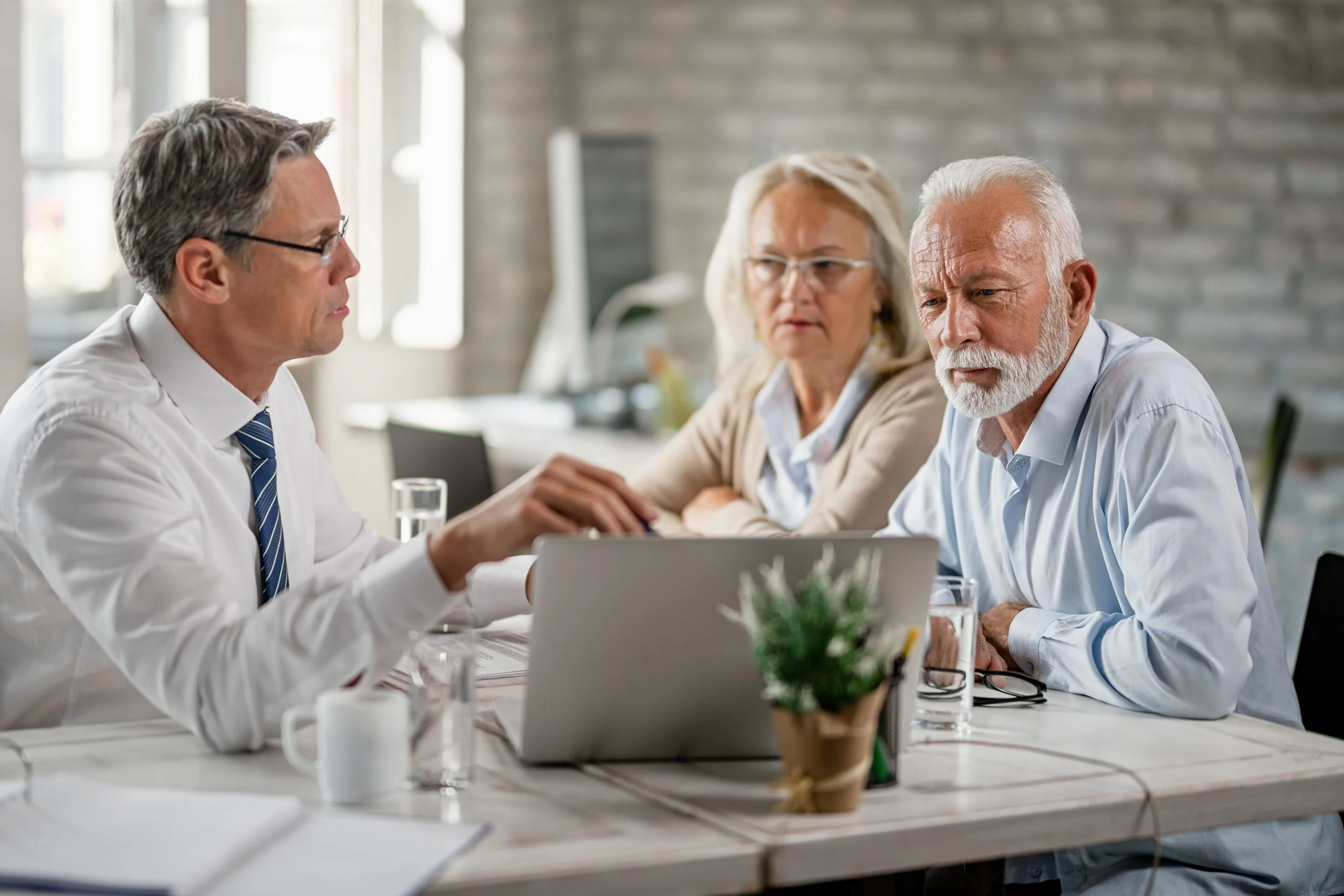 Three older adults in a meeting room, engaging in a discussion with a man in a white shirt and tie, sitting at a table with a laptop, glasses, and a potted plant.