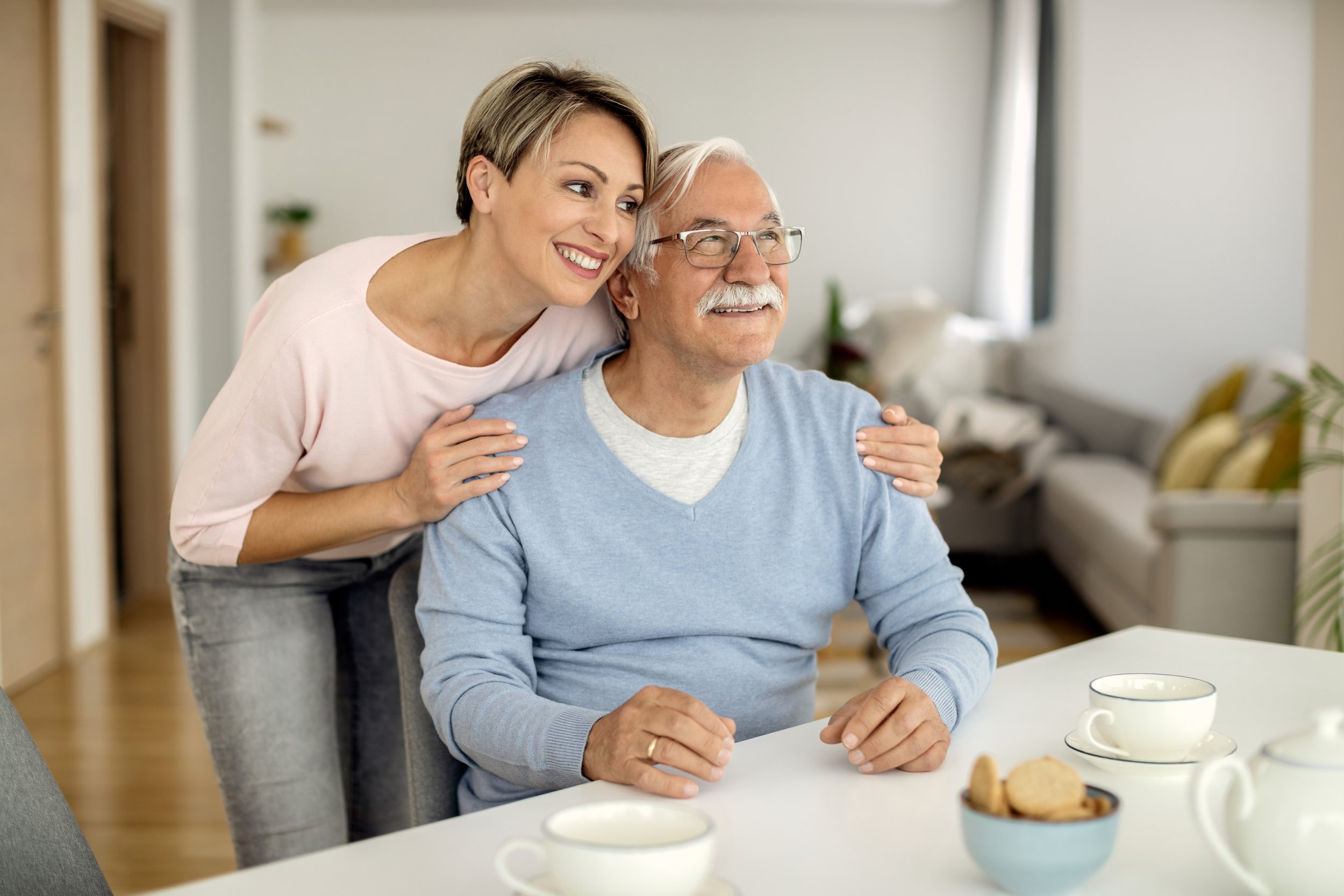 An older man wearing glasses and a light blue sweater is smiling while sitting at a table with tea cups and cookies. A woman with short blonde hair, wearing a pink top, is standing behind him and softly holding his shoulders, smiling warmly.