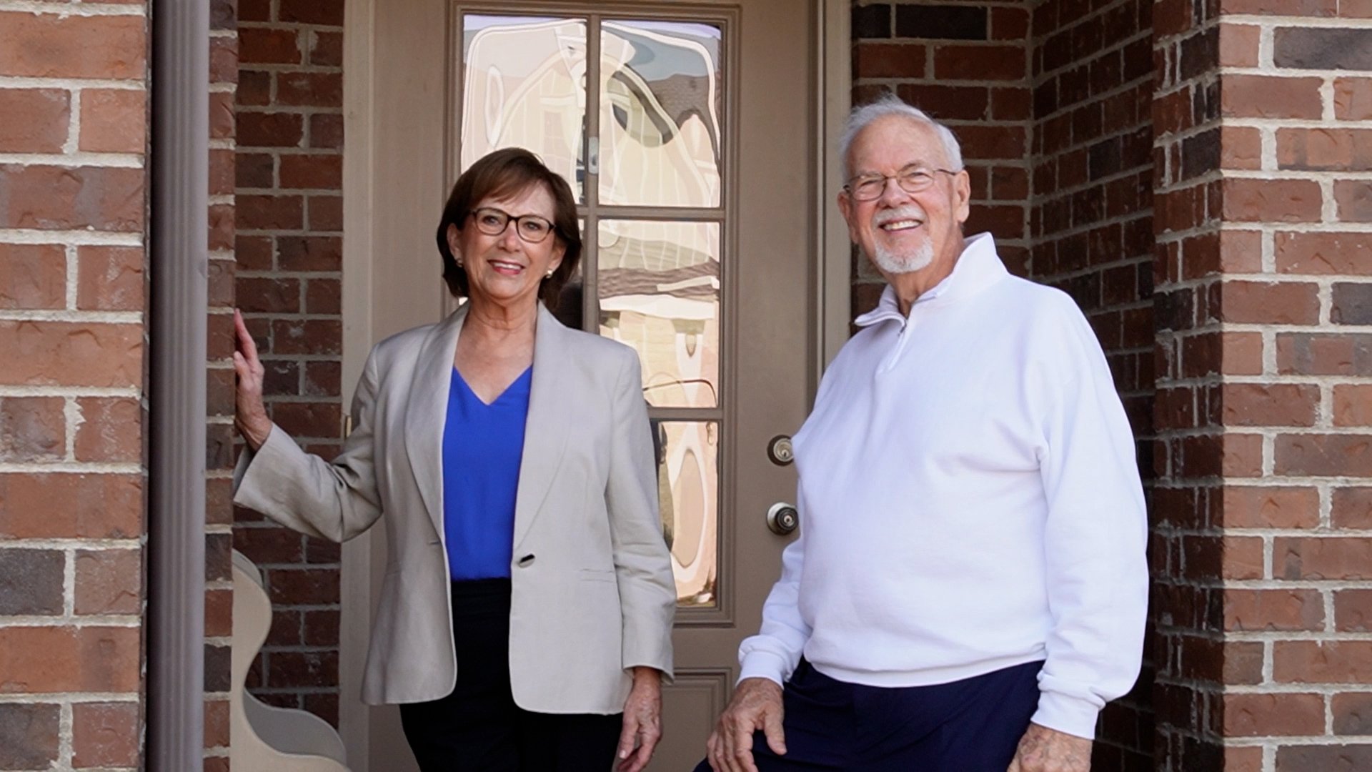 A smiling elderly woman wearing glasses and a beige blazer standing next to a man in a white sweater outside a house with a brick exterior, a door with a glass window, during daytime.