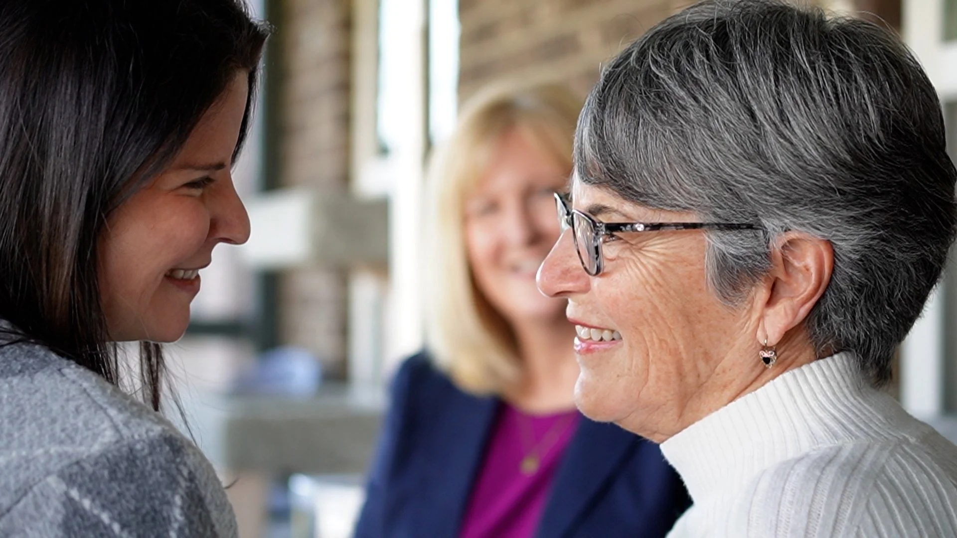 Three women smiling at each other indoors, with one woman in focus wearing glasses and a white sweater, the other two women slightly blurred in the background, engaged in a friendly interaction.