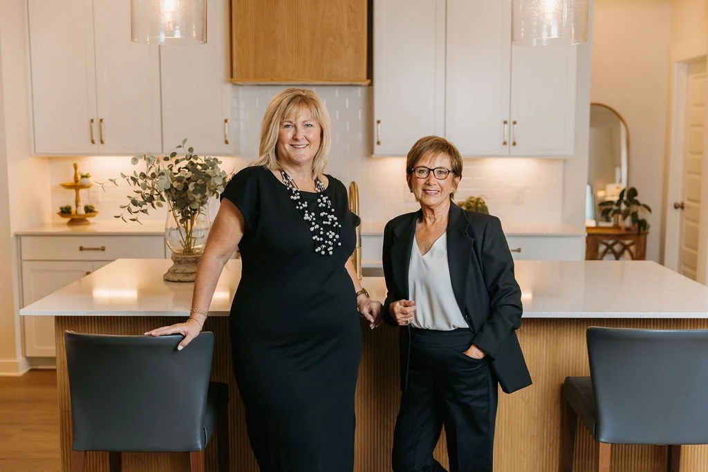 Two women standing in a modern kitchen, smiling at the camera. One woman is wearing a black dress with jewelry, and the other woman is dressed in a dark blazer and glasses.