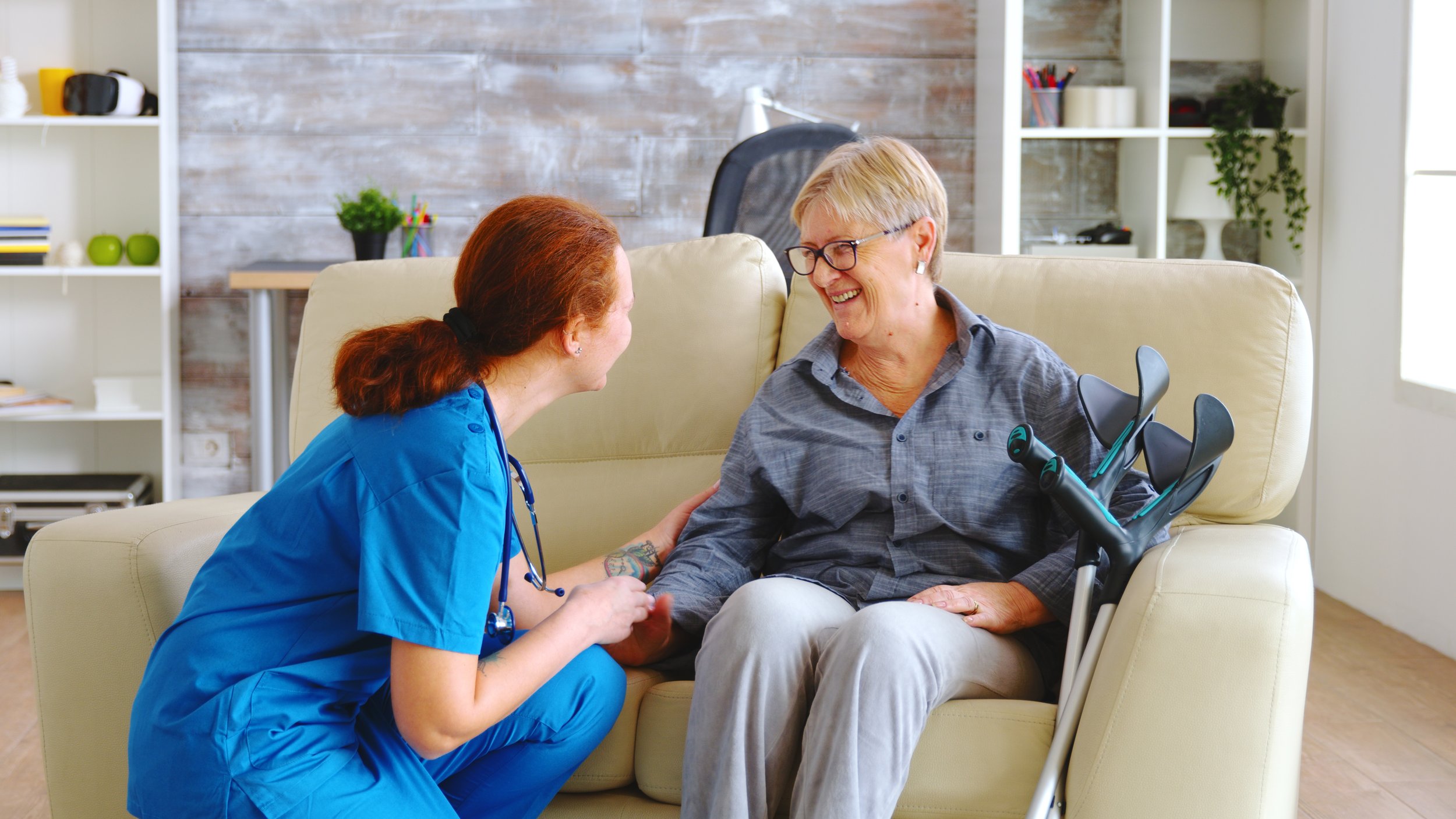 A healthcare worker in blue scrubs and a stethoscope talking to an elderly woman sitting on a cream sofa, smiling and holding the woman's hand in a cozy, well-lit room.
