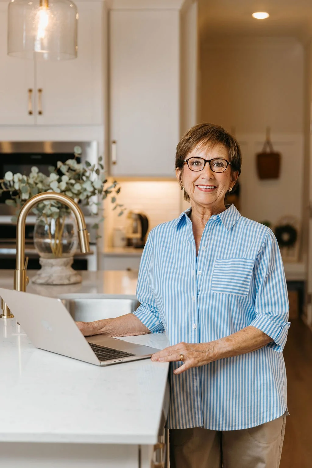 An elderly woman with short brown hair, glasses, wearing a blue and white striped shirt, stands in a modern kitchen, smiling at the camera with a laptop on the counter in front of her.