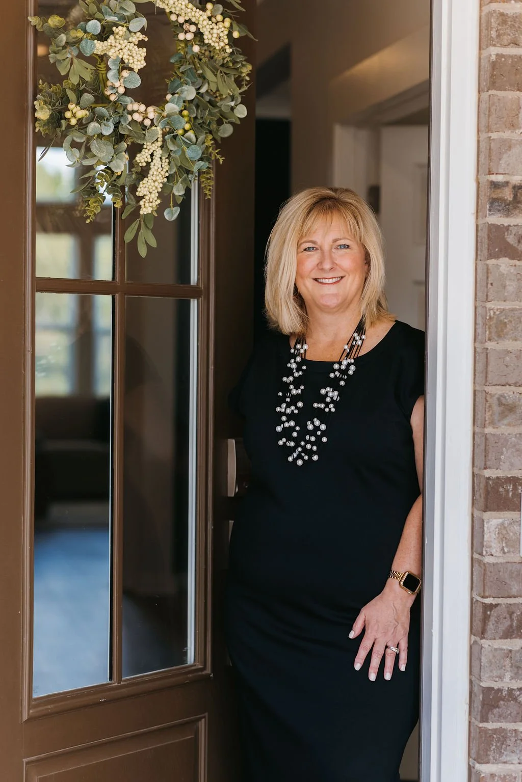 A woman in a black dress smiling, standing at an open door with brick wall on the side, and a floral decoration hanging above.