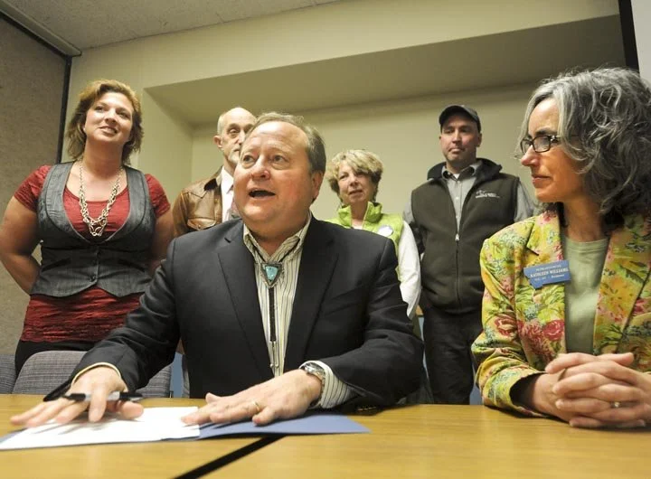 Six people gathered around a man in a suit sitting at a table, appearing to be at a formal meeting or event, with three women and two men standing behind him.