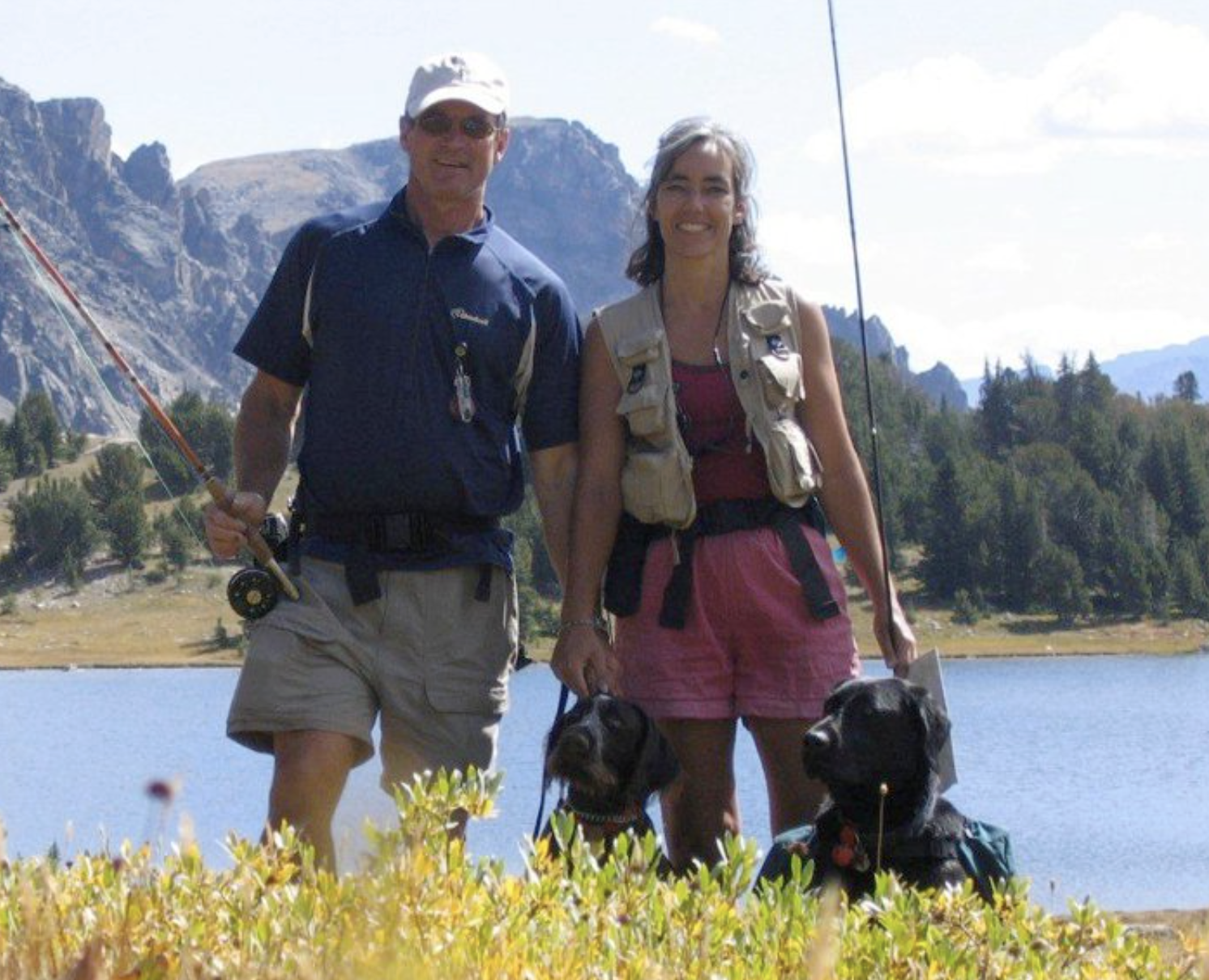 A man and a woman standing outdoors near a lake, holding two dogs, with mountains and trees in the background during daytime.