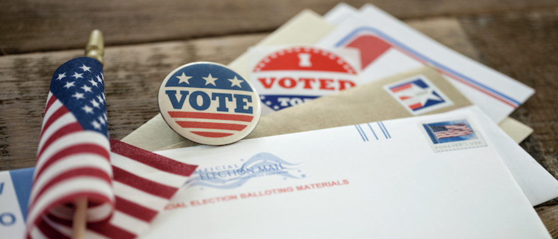 American flag and 'Vote' stickers, along with election mail-in ballot materials on a wooden surface.