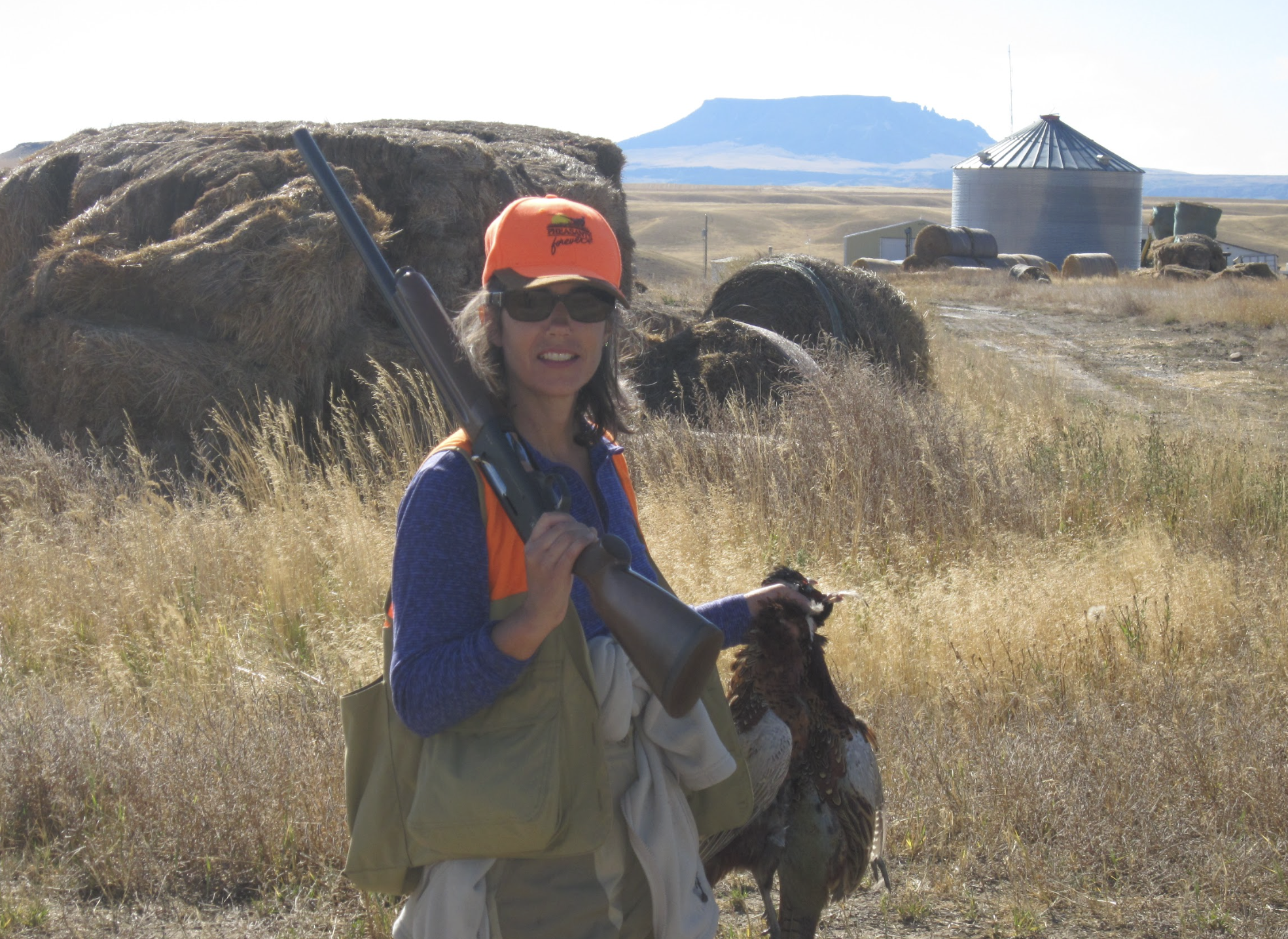 Kathleen Williams hunting, holding a rifle, standing in a grassy field with hay bales and a turkey at her feet. She is wearing an orange cap and sunglasses, with a blue jacket and tan vest.