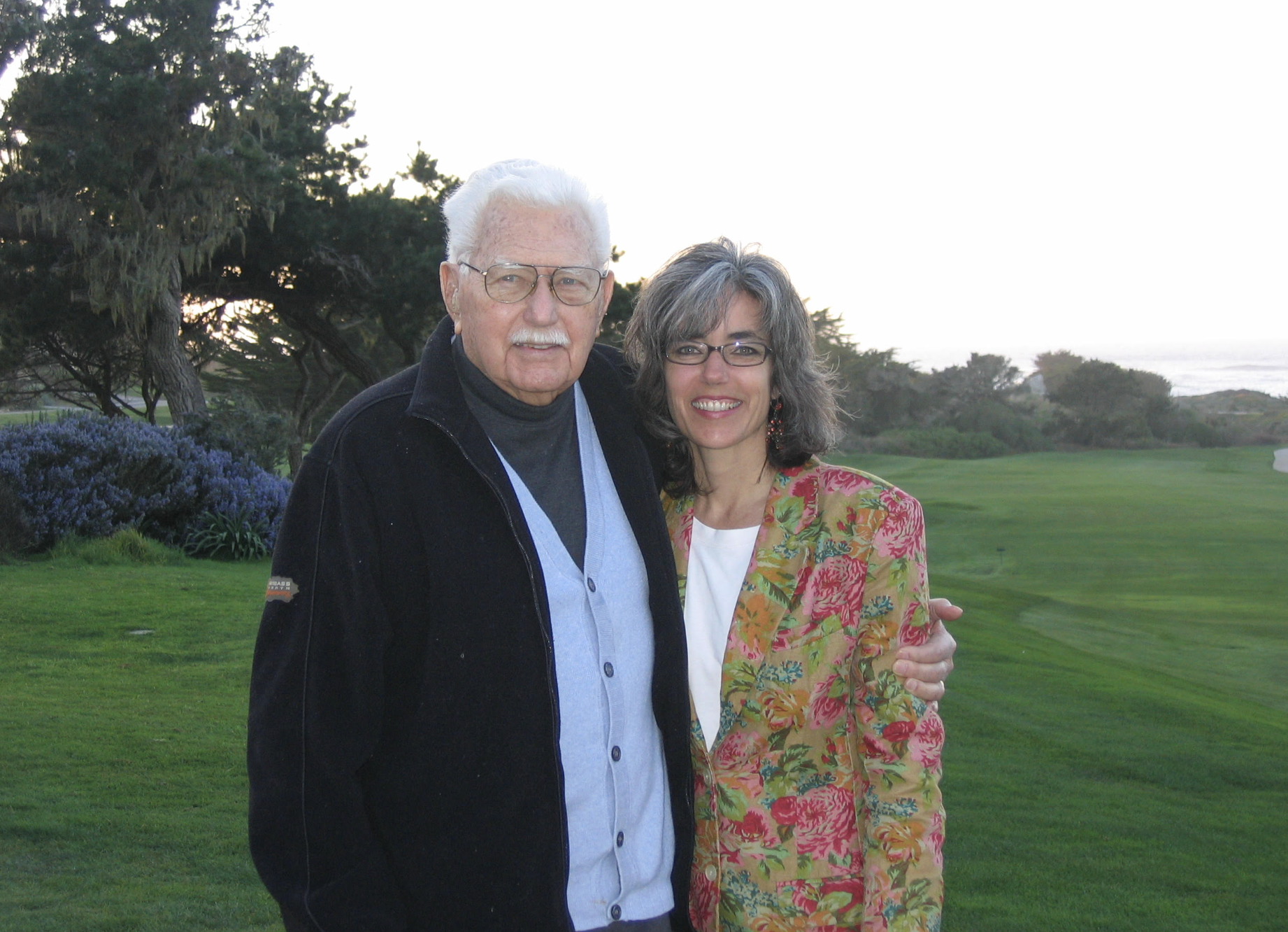 A middle-aged man with white hair, glasses, and a dark jacket stands beside a woman with gray hair, glasses, and a colorful floral blazer. They are outdoors on a grassy area with trees and shrubs, possibly a golf course, with the ocean in the background.
