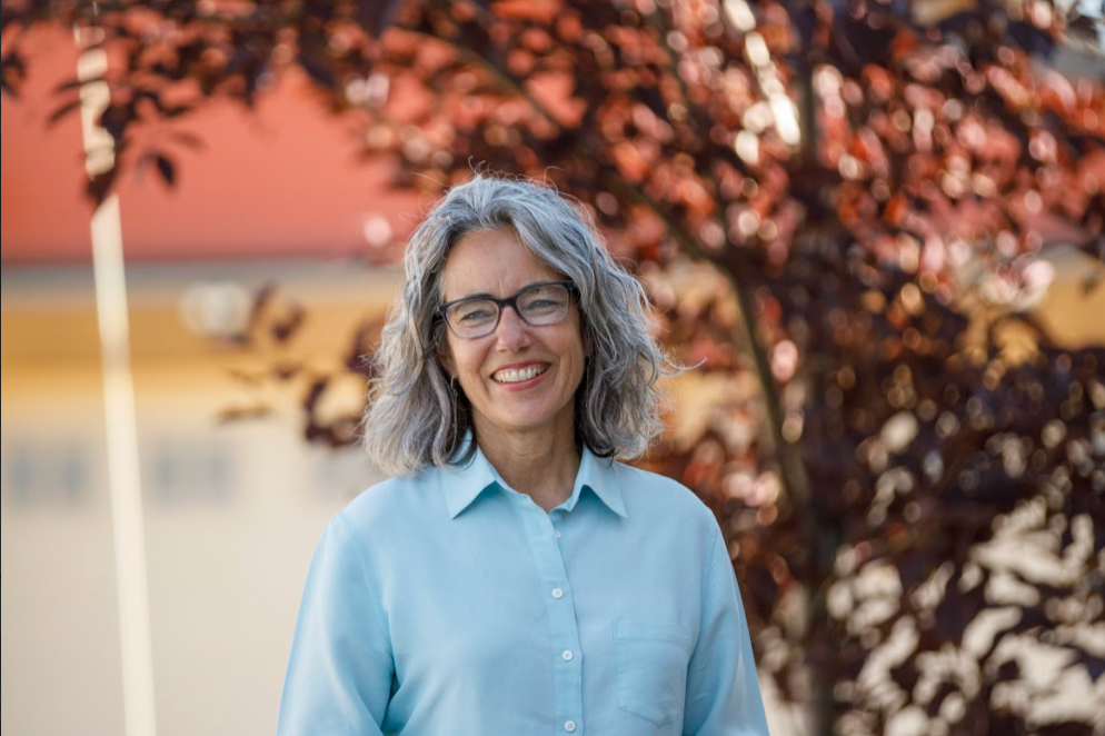A smiling middle-aged woman with gray hair and glasses standing outdoors in front of a tree with reddish leaves.