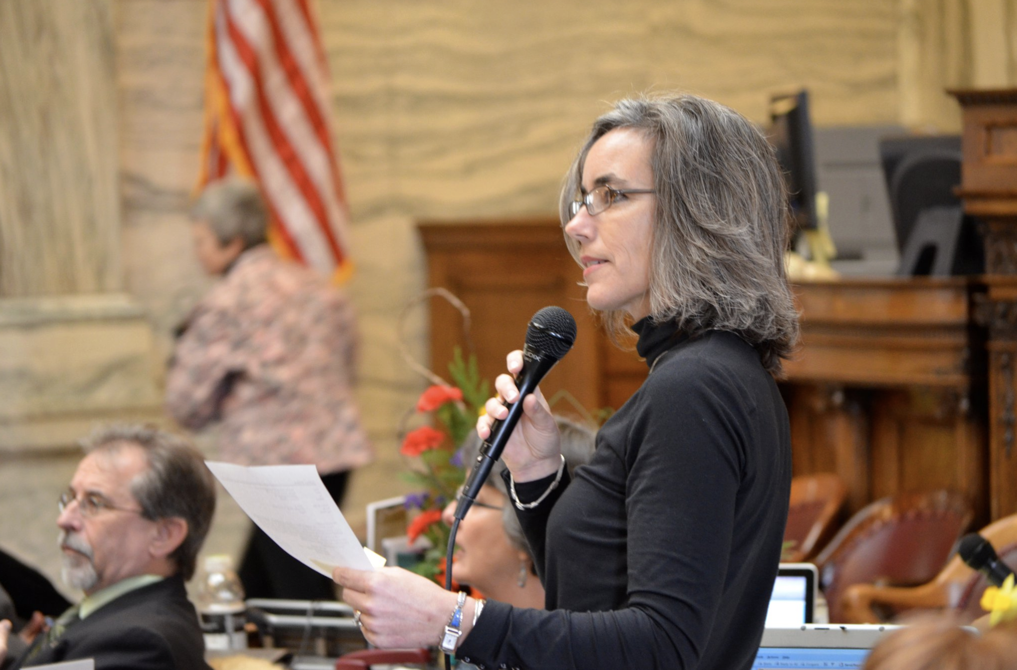 A woman with glasses holding a microphone and reading from a piece of paper at a meeting or conference, with an American flag and other attendees visible in the background.