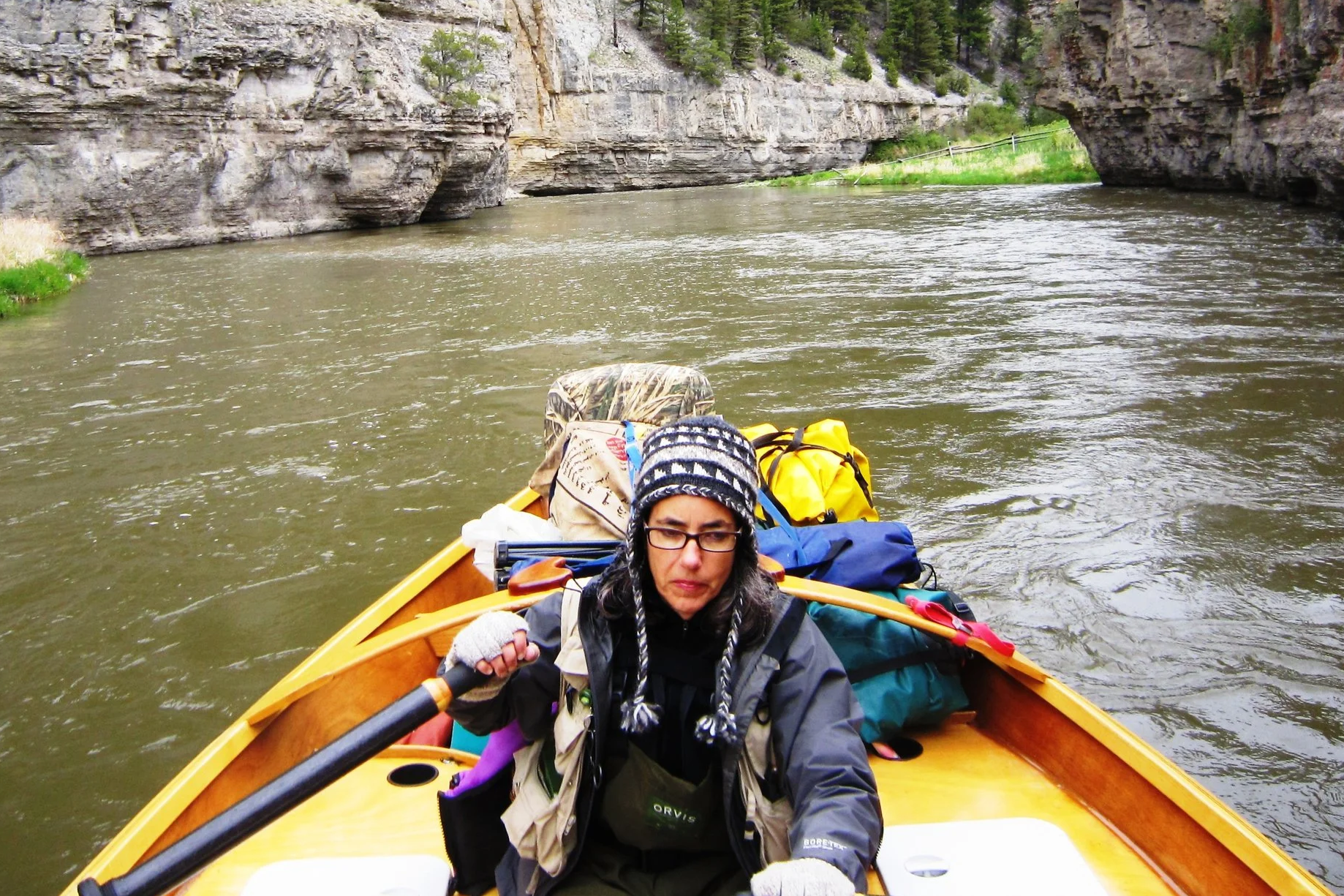 Woman wearing a knit cap and glasses paddling a yellow boat on a river surrounded by rocky cliffs and green trees.