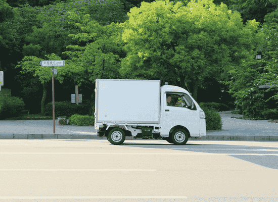 Small white delivery truck driving recycled glass on a city street with green trees and a sidewalk in the background.