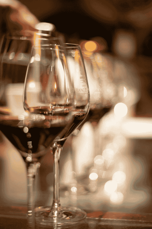 Glasses of red wine are lined up on a warmly lit bar