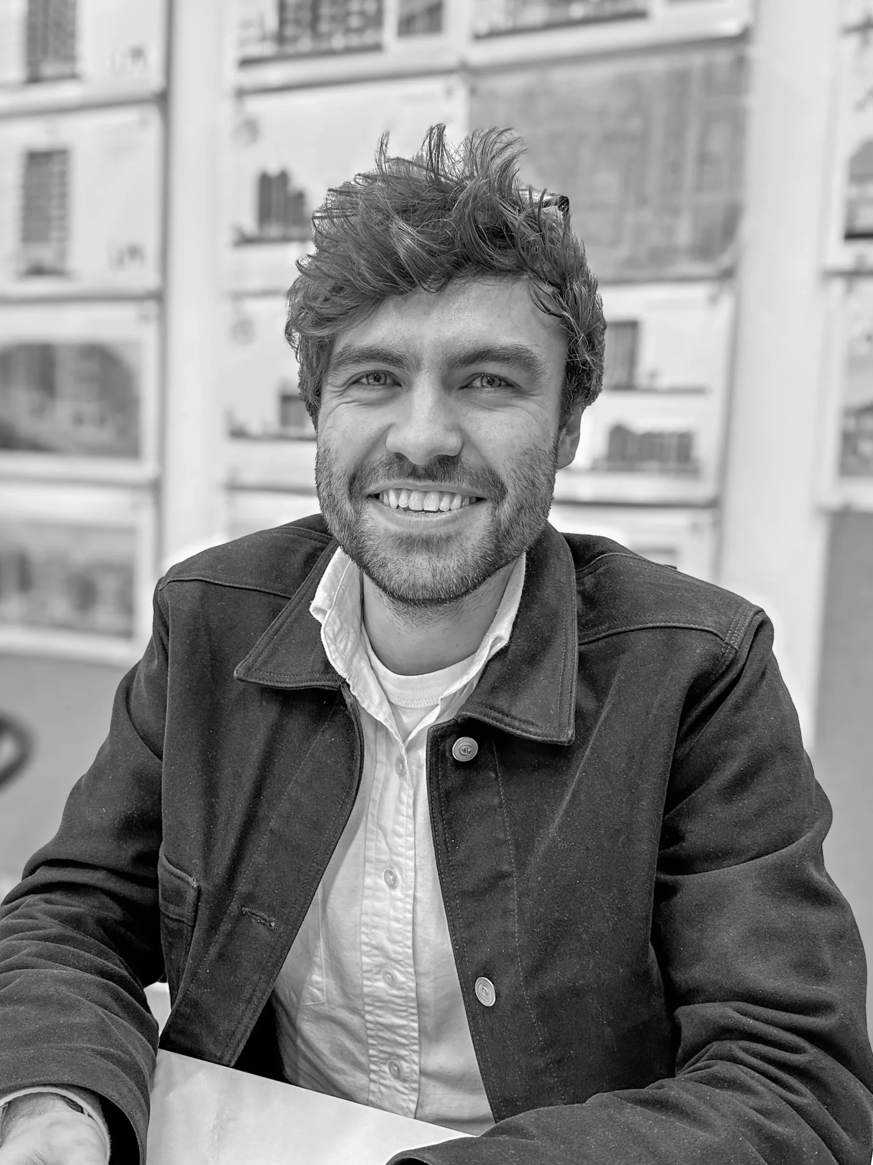 A smiling young man with tousled hair and a beard, wearing a white shirt and a dark jacket, sitting at a table with a blurred background of windows and buildings in black and white.