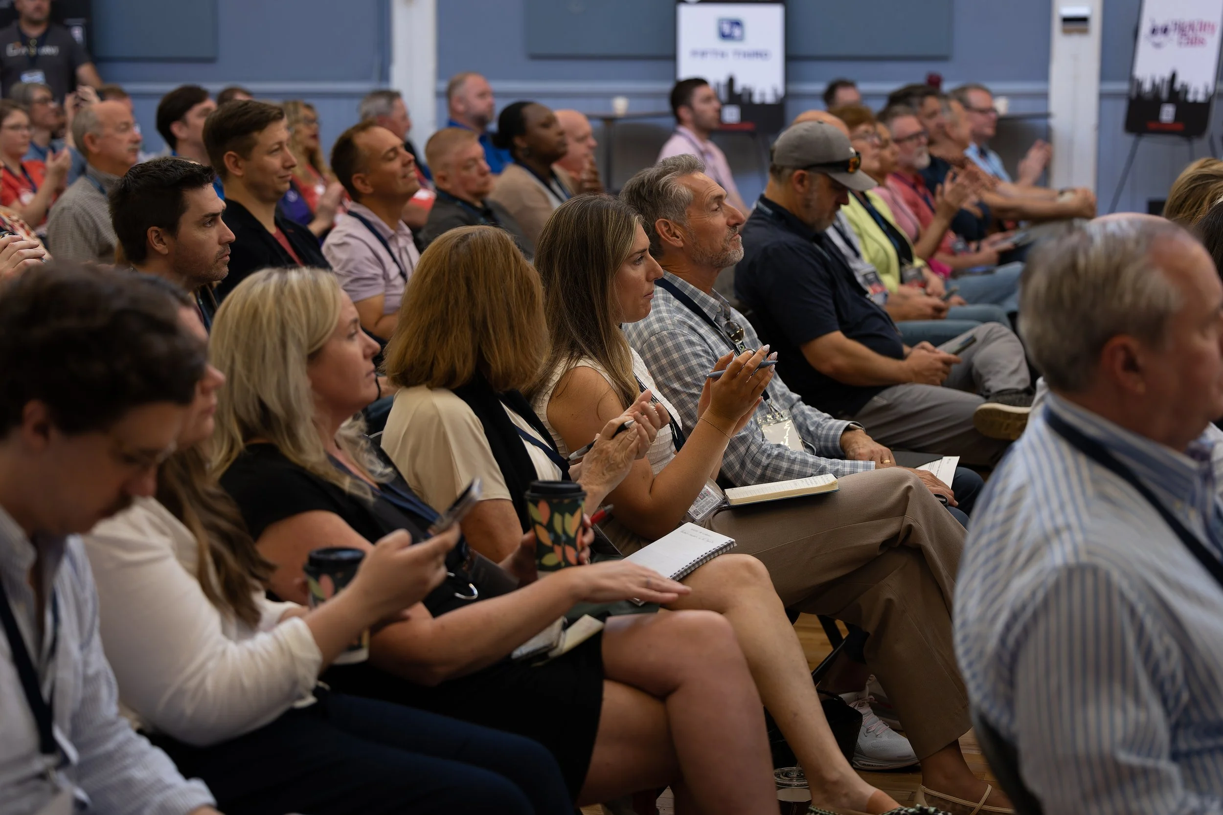Audience seated in rows at a conference, some taking notes, some using smartphones.
