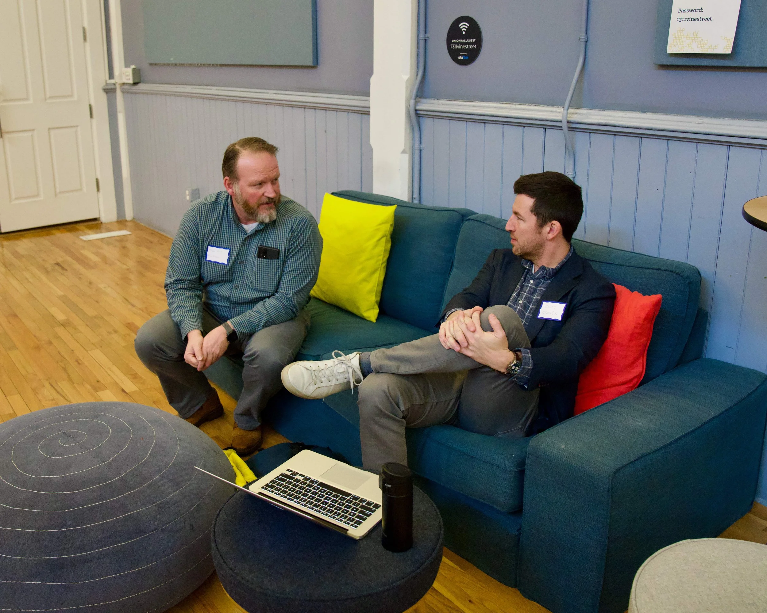 Two men sitting and talking in a office lounge. One is sitting on a blue couch with a yellow pillow, and the other is sitting on a black pouf. A laptop and a black water bottle are on a small round table in front of them.