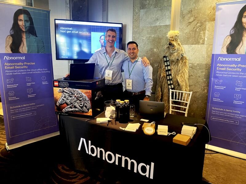 Two men standing behind a table at a booth for Abnormal, a cybersecurity company, with banners on each side and a Darth Vader LEGO model in front and a Chewbacca costume character to the right.