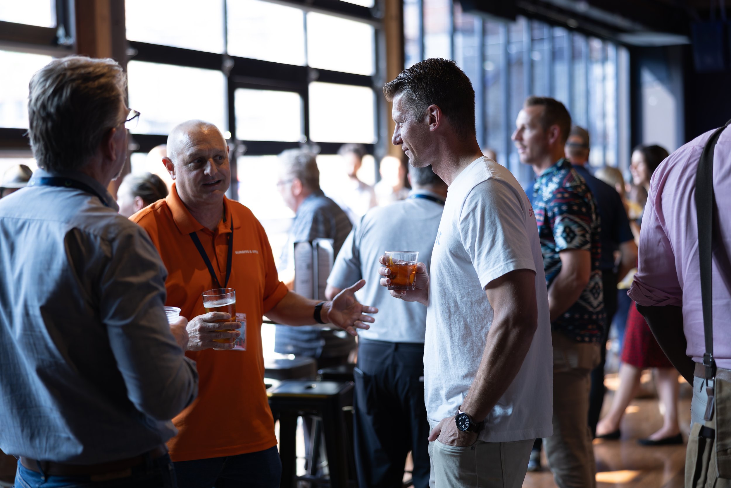 People at a networking event, engaged in conversation, standing indoors with large windows, some holding drinks.