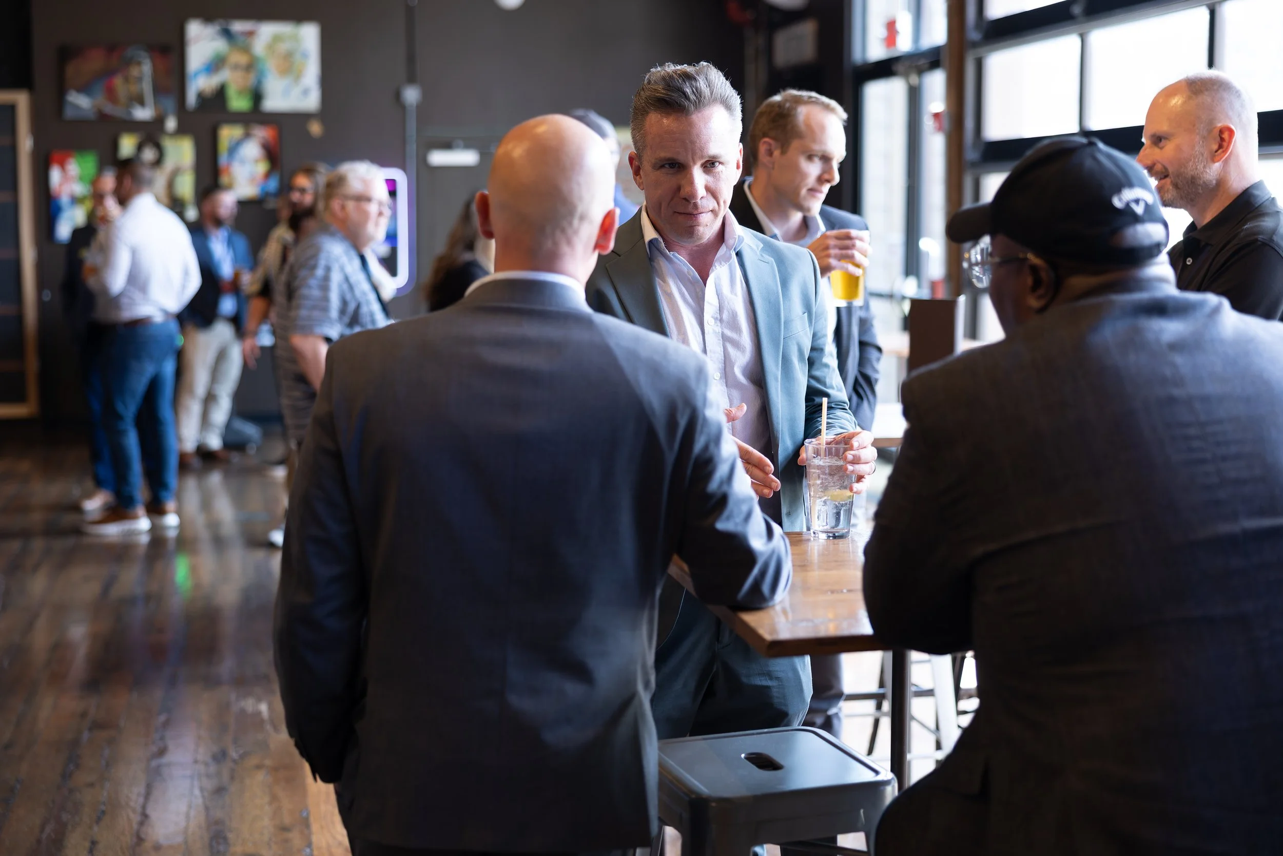 Group of people socializing in a modern bar or café, some holding drinks, with artwork on the walls and large windows letting in natural light.