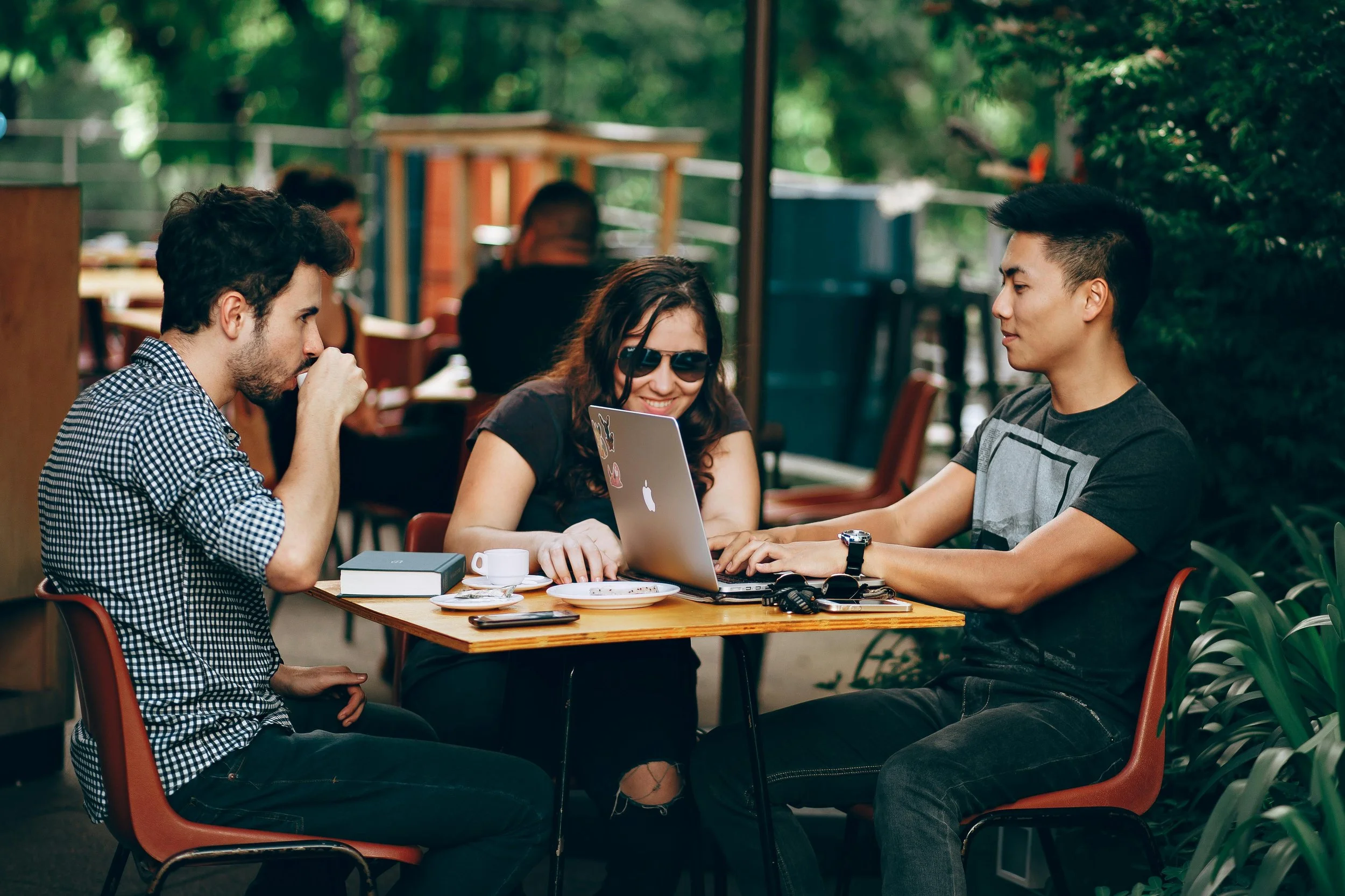 Three friends sitting at a wooden table outdoors, with one person using a laptop, one wearing sunglasses, and the third drinking from a cup.
