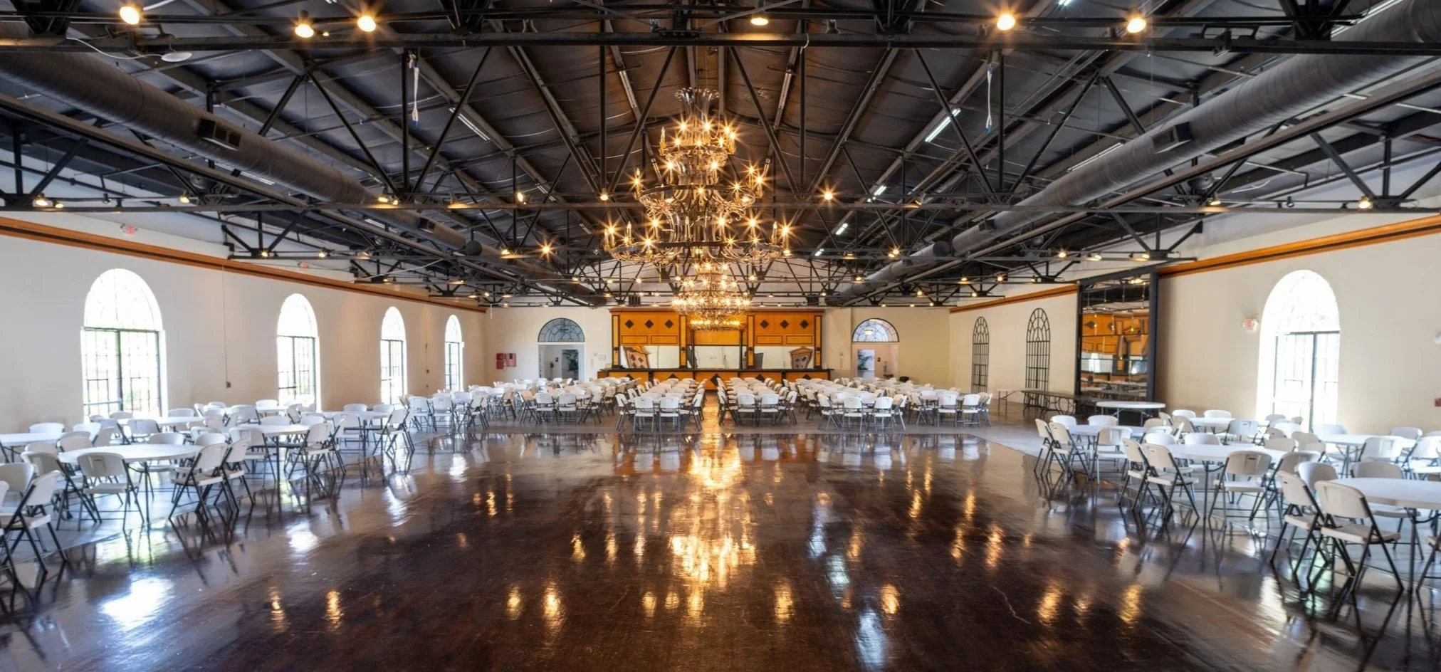 Empty banquet hall with multiple round tables and white chairs, a polished wooden floor, large arched windows, and a grand chandelier hanging from the ceiling. The room is decorated in a classic style with a stage at the front.