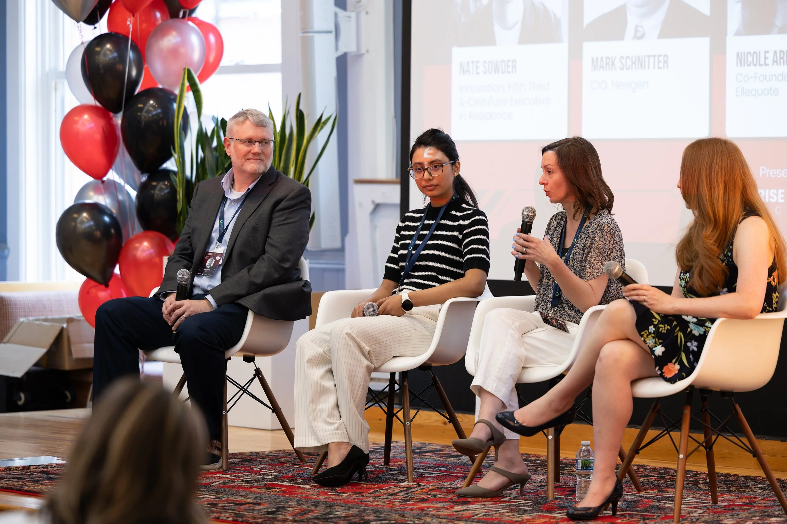 A panel discussion at a conference, featuring four diverse speakers sitting in white chairs, holding microphones. The background has a projection screen displaying names and titles, with red, black, and pink balloons on the left side and a plant behind the speakers.