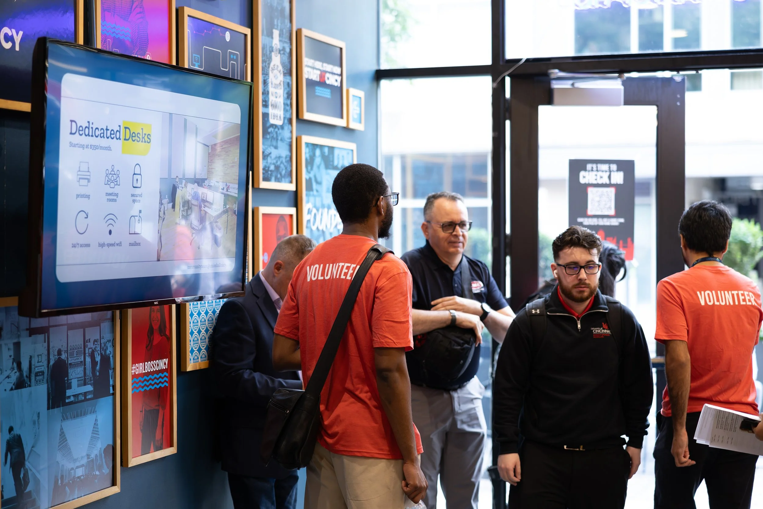 People waiting inside a building near the entrance, some wearing orange volunteer shirts, with a large screen displaying amenities and a QR code check-in sign at the door.