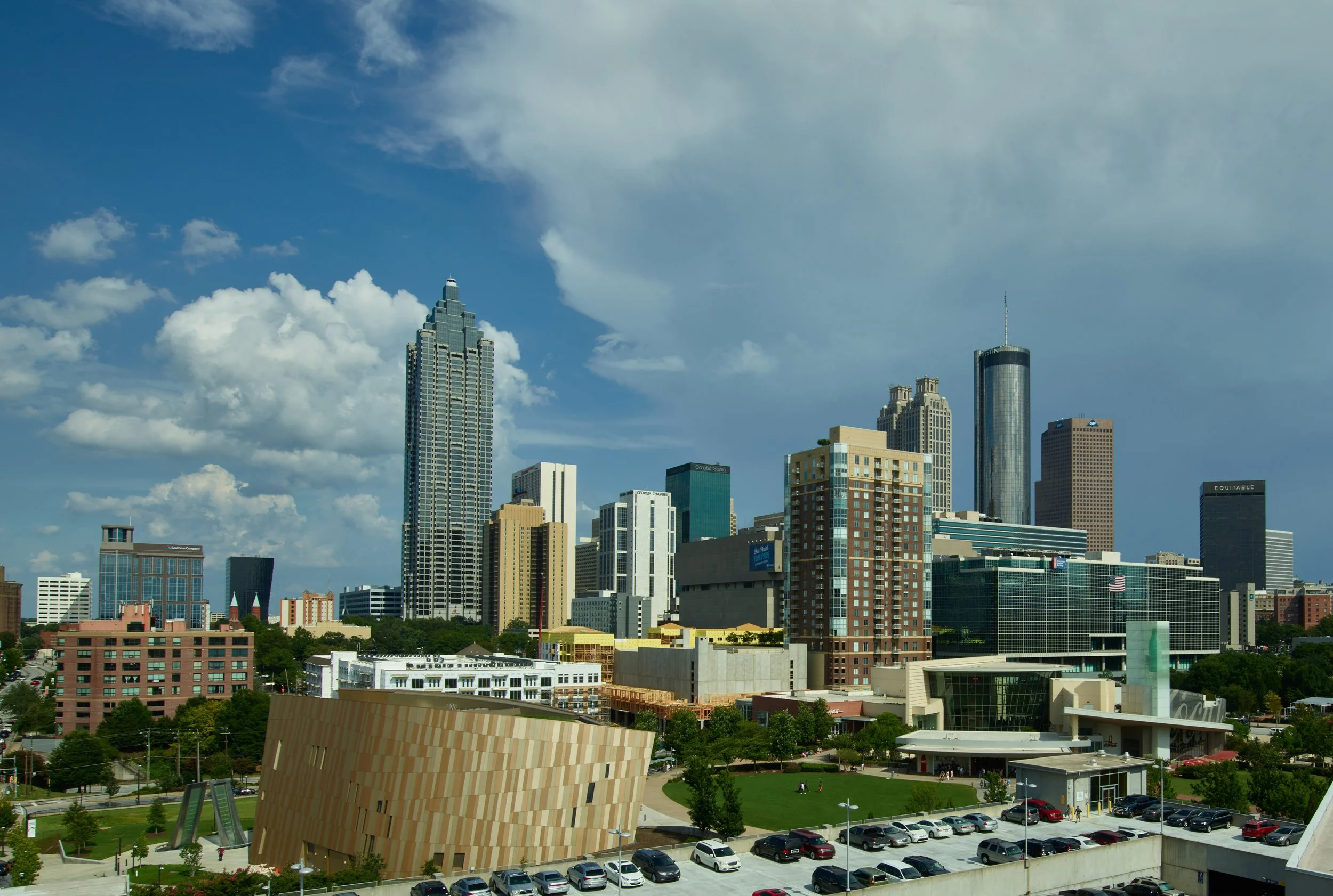 Skyline of a modern city with skyscrapers, a blue sky, and some clouds. Green park and parking lot visible in the foreground.