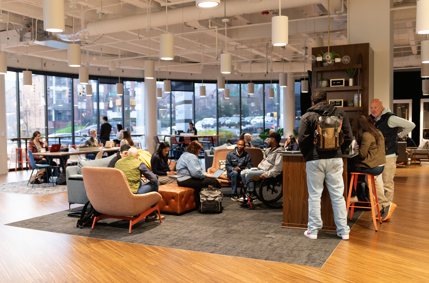 People gathered in a modern, well-lit lobby or lounge area with large windows. Some are sitting on sofas and chairs, working on laptops or having conversations. A small group stands at a bar-height counter, talking. The space has contemporary furniture, wooden floors, and decorative shelves.