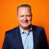 A blurred portrait of a person with short dark hair wearing a black top against a light background.