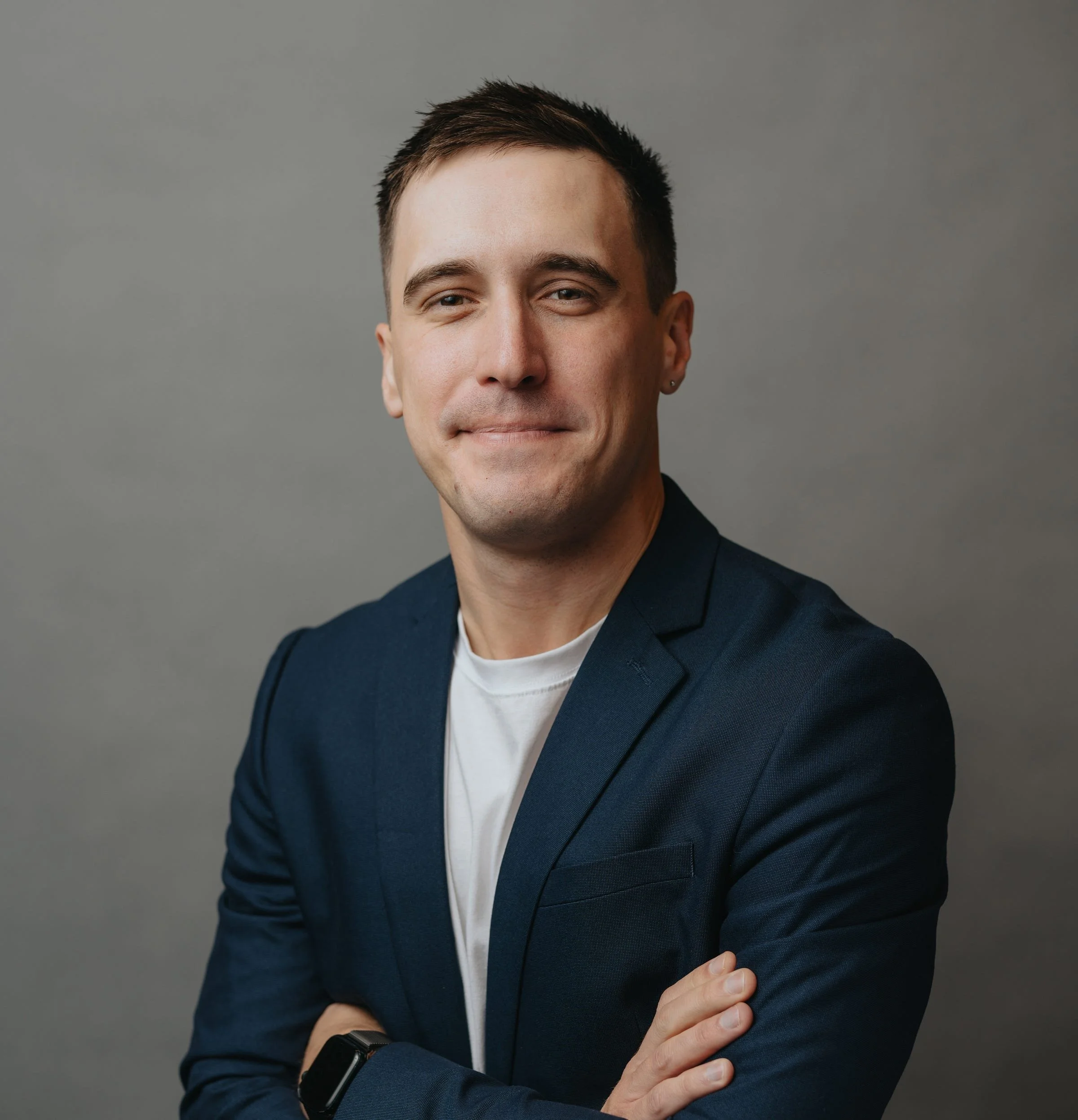 Professional man with short brown hair, wearing a navy blazer and white t-shirt, crossing arms, smiling slightly, against gray background.