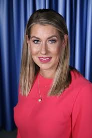 Blurred image of a person with short dark hair, wearing a black top, against a light background.