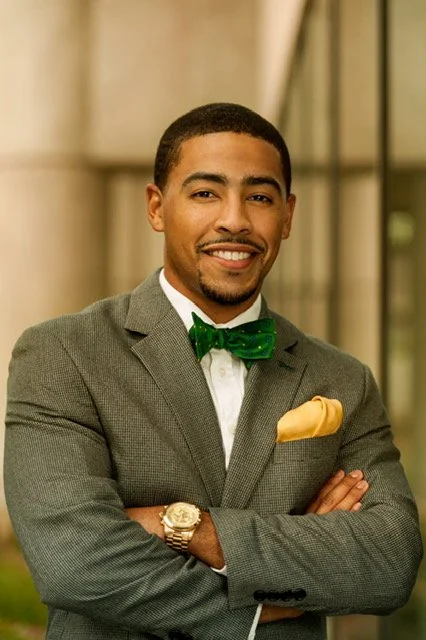Young man in gray suit with green bow tie and yellow pocket square, smiling with arms crossed in an urban setting.